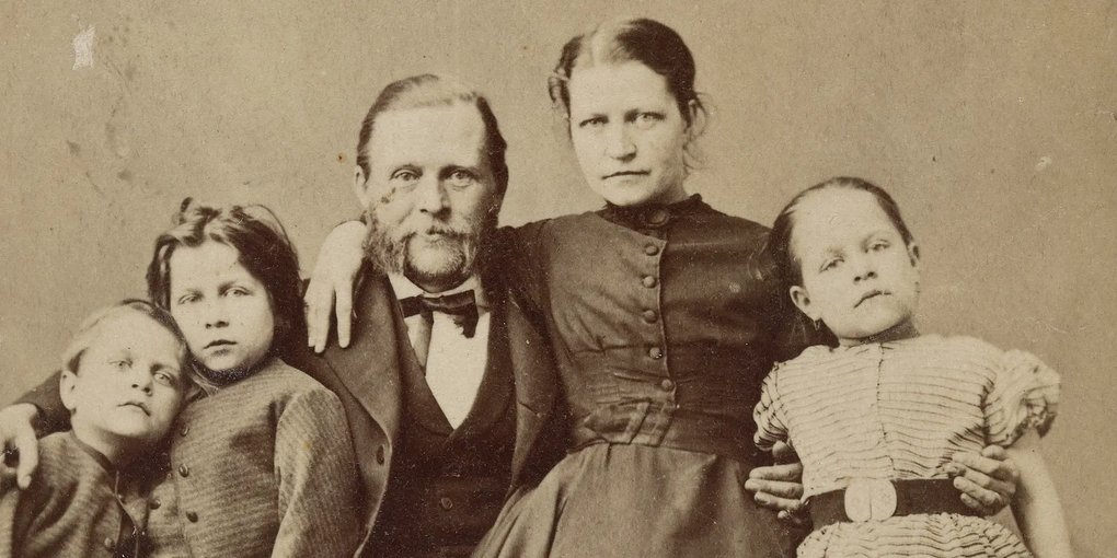 An old black-and-white family portrait shows two adults and three children gathered closely together, facing the camera. They wear period clothing and have serious expressions. The image conveys a formal and quiet atmosphere from a bygone era.